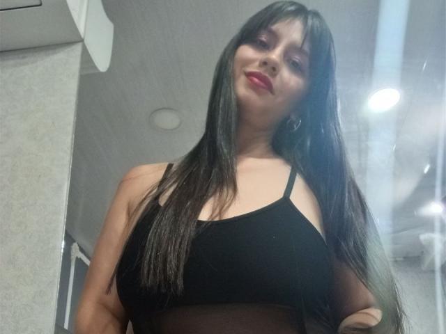 MadissonFontaine with long black hair, wearing a black bralette, showing her big tits, red lipstick, and a slight smile, standing in a dimly lit room.