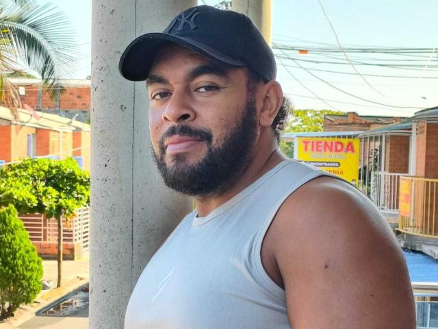 JordanoSexHot, musculoso, com barba, pele bronzeada, vestindo um boné preto e uma camiseta branca sem mangas, em pé contra um poste cinza, sorrindo levemente, com uma palmeira ao fundo..