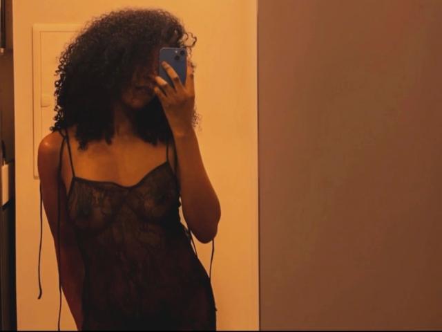 Sexyblackqueen-sensuelle, avec des cheveux bouclés, portant une nuisette noire en dentelle transparente, montrant de petites seins et des mamelons, prenant un selfie devant un miroir avec un téléphone bleu dans une pièce peu éclairée..