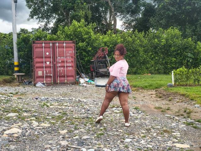 YiyaHot69, curvy with big ass, wearing a pink top and floral skirt, standing on rocky ground, looking back, red cargo containers and greenery in the background.
