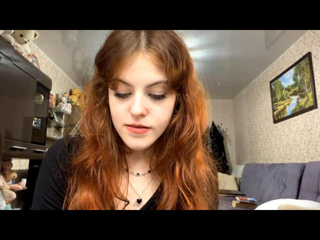 ArvillaUptmor a les cheveux roux et ondulés, une peau pâle et un eye-liner foncé. Elle regarde vers le bas avec une légère moue, portant un haut noir et un collier avec un petit pendentif noir..