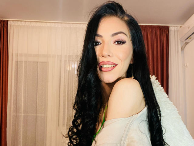 SinReea with long black hair, red lipstick, and a smile, showing her shoulder and off-shoulder top, looking at the camera, has cute tits, and is in a cozy room.