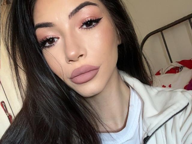 SinReea has dark, straight hair, light skin, and full pinkish-brown lips with thick eyeliner. She wears a white shirt and a white jacket, looking serious.