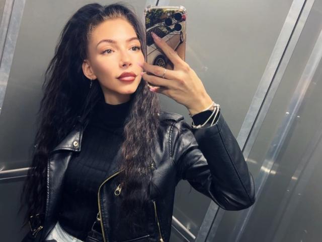 SinReea with long black hair, wearing a black leather jacket and black ribbed turtleneck, taking a selfie in an elevator, showing off her smooth, cute face and red lipstick.