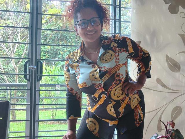 YiyaHot69 with curly red hair, glasses, and a colorful patterned shirt, leaning on a window, smiling. She's got a cute face and a curvy ass.