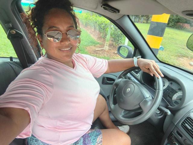 YiyaHot69 with glasses, pink shirt, and purple shorts, sitting in a car, smiling, right hand on the steering wheel, left arm raised, showing a cute, chubby ass.