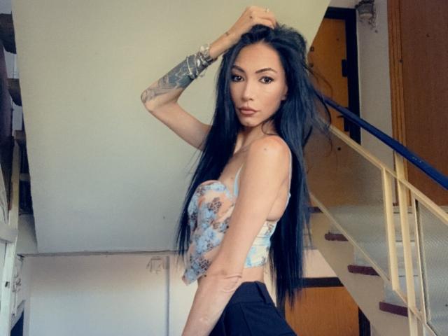 SinReea with long black hair, blue floral top, and black pants, showing tattooed arm, standing in front of stairs, looking at camera with hand in hair.