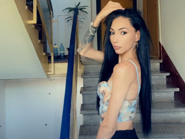 SinReea with long black hair, tattoos on her right arm, wearing a floral bra and black pants, standing on stairs, looking back, hand in hair, wristwatch visible.