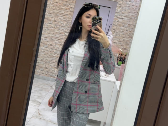 SinReea mit langen, schwarzen Haaren, die einen grauen Blazer mit Karomuster, ein weißes Oberteil und graue Hosen trägt, macht ein Selfie vor einem Spiegel mit ihrem Handy. Sie trägt Ohrringe mit Kreuzen und eine Sonnenbrille..