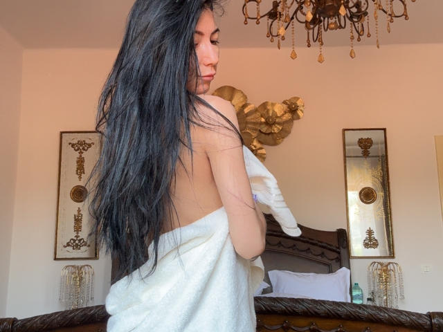 SinReea with long black hair, back to camera, wrapped in a white towel, holding a glove, standing in a room with a chandelier and decorative wall pieces.