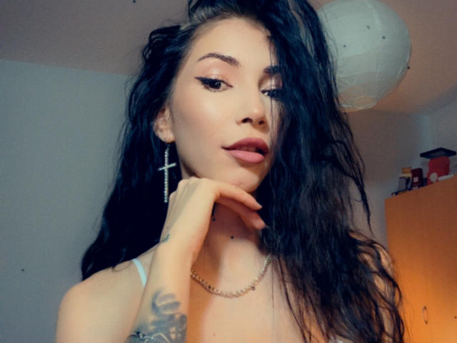 SinReea, con i suoi lunghi capelli neri e ondulati, indossa orecchini a forma di croce e una collana, ha una mano sul mento, mostrando un tatuaggio e labbra piene, in un selfie..