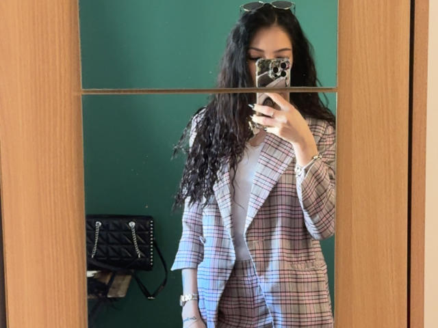 SinReea mit welligem, schwarzem Haar, die einen karierten Blazer über einem weißen Hemd trägt und ein Handy mit einer Handyhülle mit Leopardenmuster hält, wobei sich das Spiegelbild in einem Spiegel zeigt..