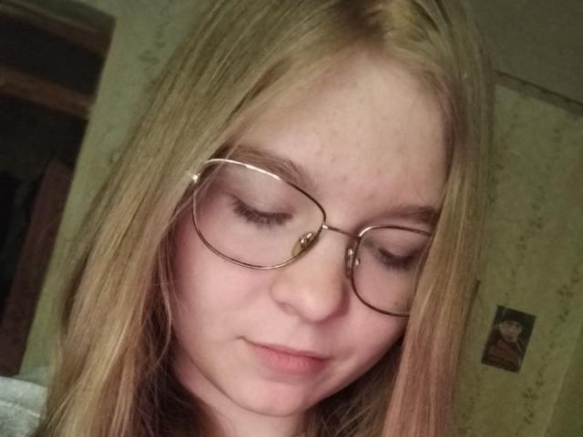 DrucillaSchab hat hellblondes Haar, trägt eine Brille und hat die Augen geschlossen, mit einem leichten Lächeln. Ihre Nase ist klein und ihre Haut ist blass..