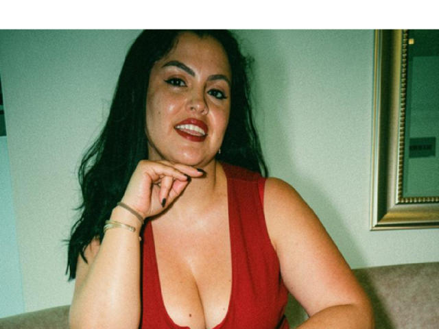 Charlotte Devereaux, met donker, golvend haar, een rode jurk, die grote borsten laat zien, lachend, met haar kin op haar hand, rode lippenstift, een groene muur, een spiegel met een gouden lijst, een beige bank, een armband om haar pols..