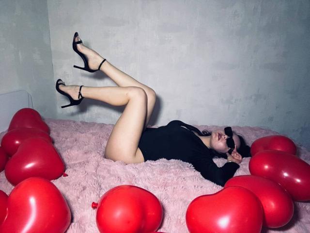 JasmineAsha, allongée sur un lit rose et moelleux, les jambes en l'air, portant des talons hauts noirs et un body noir, entourée de ballons en forme de cœur rouges, avec des lunettes de soleil noires..