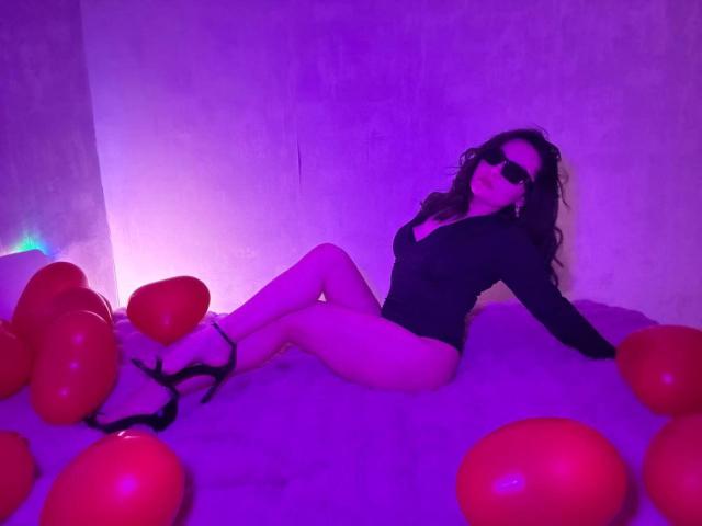 JasmineAsha, avec ses cheveux noirs bouclés, ses lunettes de soleil noires, son haut noir à manches longues et ses talons hauts, assise sur un lit violet avec des ballons rouges, montrant ses fesses et ses jambes..