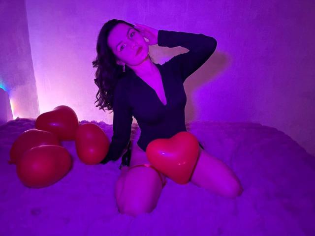 JasmineAsha, avec ses cheveux bruns bouclés, portant un haut noir à manches longues, agenouillée sur un lit rose, entourée de ballons en forme de cœur rouges, sous un éclairage violet, touchant son visage..