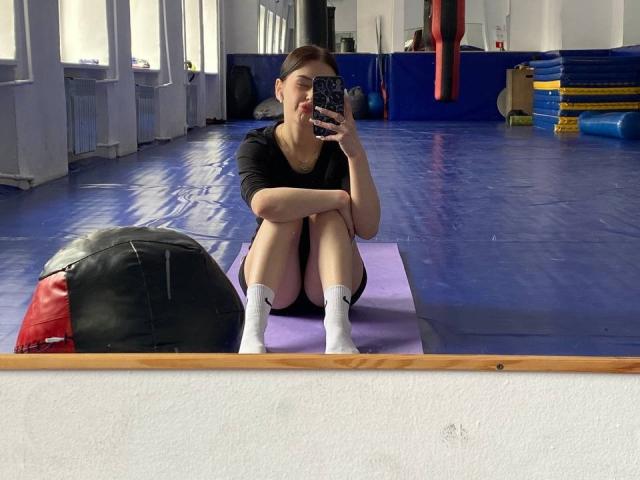 LydiaSween zit op een paarse mat, met gebogen benen en gekruiste armen, gekleed in een zwarte top, witte sokken en zwarte korte broek, en maakt een spiegelzelfportret in een sportschool..