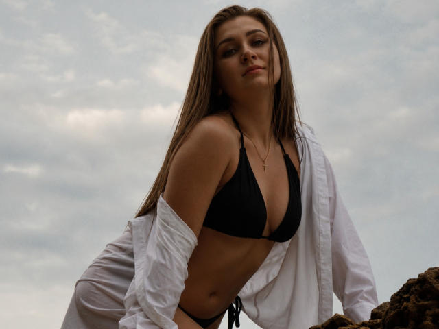BeataAngel con lunghi capelli castani, indossa un top da bikini nero, una camicia bianca portata a spalle scoperte, appoggiata a delle rocce, cielo nuvoloso, una piccola collana d'oro, guarda in basso, con un leggero sorriso..