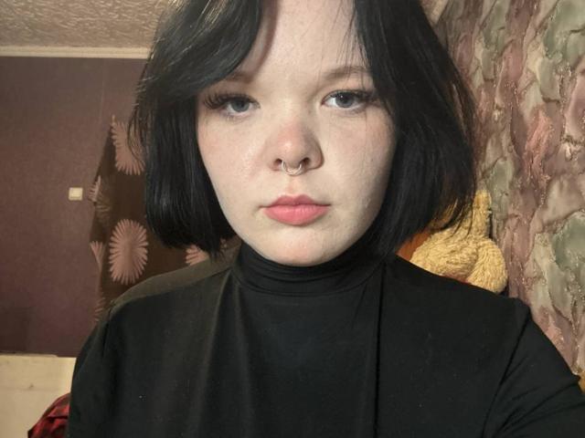 Mikaela Etherton mit heller Haut, schwarzem Bob, Nasenring und schwarzem Rollkragenpullover, wirkt ernst mit leicht geschwollenen, rosafarbenen Lippen, trägt Eyeliner, in einem schwach beleuchteten Raum..