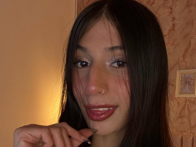 Sophie Beaumont a de longs cheveux noirs, un piercing au nez et un rouge à lèvres rose brillant. Elle sourit, ses lèvres rouges, et sa main est près de son menton. Ses yeux sont maquillés avec des paillettes..