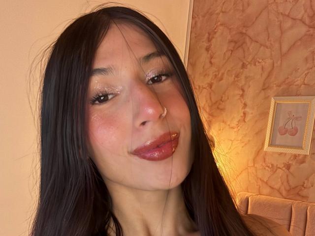 Sophie Beaumont a des yeux brillants et étincelants, un anneau de nez et des lèvres rouges et brillantes. Ses cheveux noirs sont légèrement en désordre, et elle sourit avec un regard subtil et séduisant..