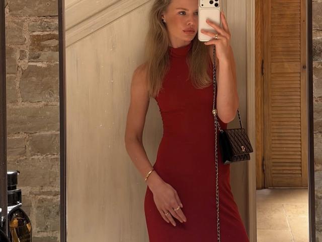 TheHottestBlonde with long blonde hair in a tight red dress, holding a phone in her right hand, wearing a black Chanel purse, standing in a mirror selfie.