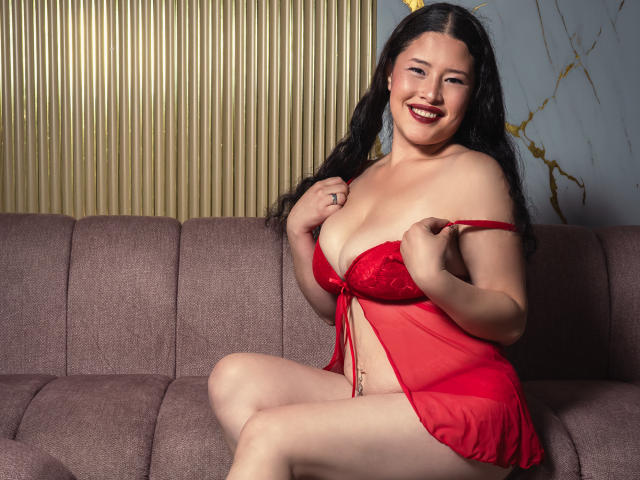 CelesteGreey está sentada em um sofá rosa, vestindo uma lingerie vermelha transparente, um modelo "babydoll" que revela seus seios e um pequeno vislumbre de sua vulva. Ela sorri com batom vermelho escuro..