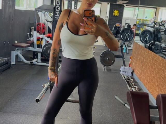 NicolSNaya with big tits, white tank top, black leggings, tattoos on arm, holding phone, standing in gym, mirror selfie, holding dumbbell, looking at phone, orange phone case.