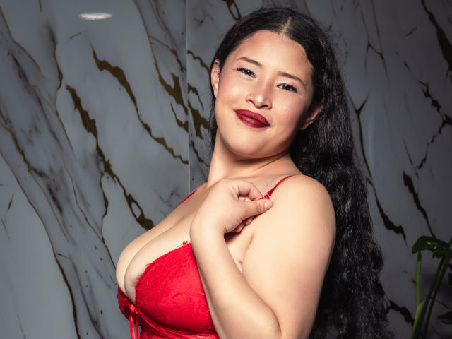 CelesteGreey mit langen schwarzen Haaren, einem roten Spitzen-BH und rotem Lippenstift, die am BH-Bügel hält, lächelnd und ihre großen Brüste zeigt, vor einer Marmorwand..