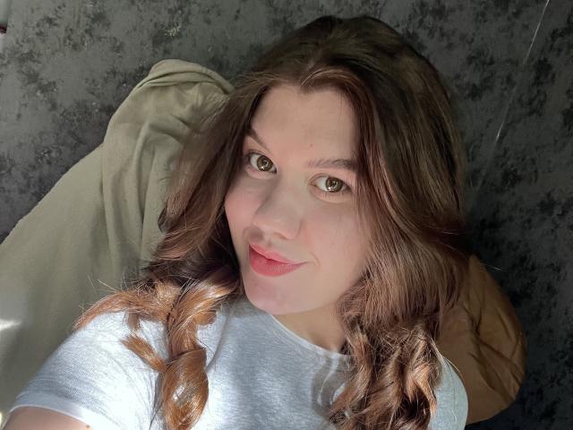 NikkiBustt, avec ses cheveux bruns clairs et ondulés, ses yeux verts et ses lèvres roses, portant une chemise blanche, allongée avec un oreiller beige, souriant à l'appareil photo..