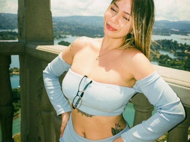 BellaDonaD mit einem hellblauen, schulterfreien Oberteil, einer Brille auf der Brust, Tattoos an der Seite, lächelnd, mit den Händen an den Hüften, auf einem Balkon, in der Sonne, mit langen, braunen Haaren, blauen Augen und heller Haut..