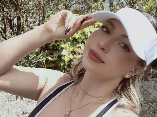 BellaDonaD with blonde hair, white cap, black tank top, and gold necklace, touching her nose with black-manicured fingers, smiling softly. Her tits are visible.