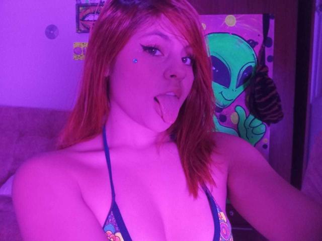 SophieHill with red hair, blue eyes, and eyeliner, sticking out her tongue, wearing a dark blue bikini top, purple-lit room, and a green alien poster in the background.