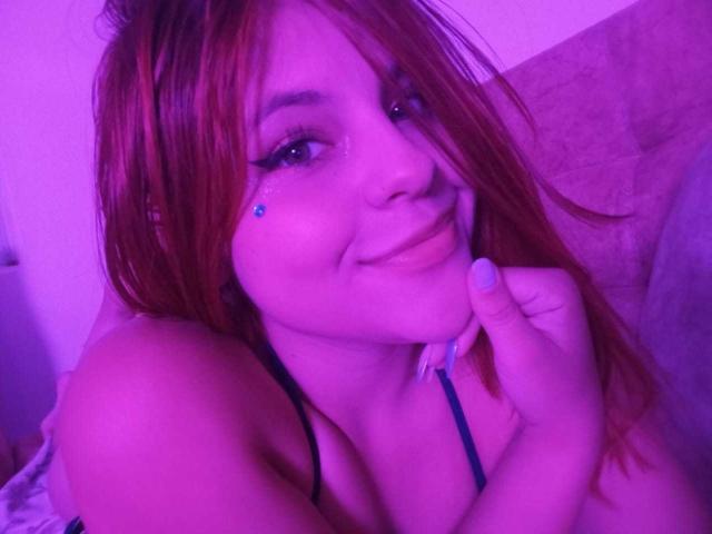 SophieHill with red hair, blue eye makeup, and a cute smile, touching her chin with her hand, showing her tits, under purple lighting.