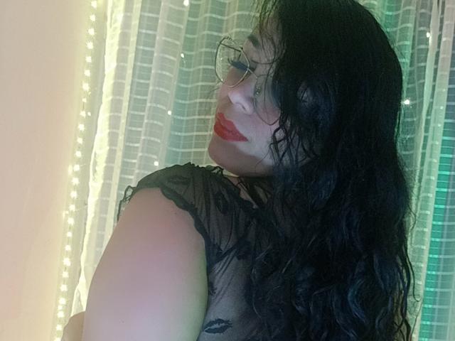 FetishLoverAna, con cabello negro ondulado, lápiz labial rojo y gafas, vestida con una blusa negra transparente, mirando por encima del hombro, mostrando su trasero, en una habitación con iluminación suave y un tono verdoso..