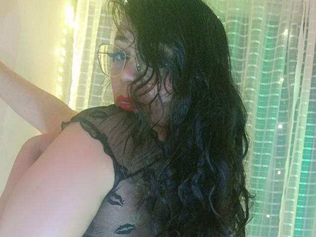 FetishLoverAna met lang, zwart, golvend haar, die een bril draagt en een doorschijnende zwarte top aanheeft, waarbij haar borsten zichtbaar zijn, rode lippenstift en een groen licht op de achtergrond..