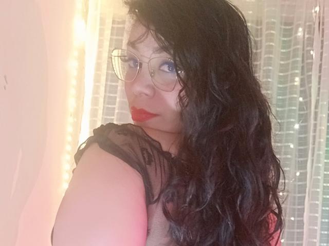 FetishLoverAna, con cabello negro ondulado, lápiz labial rojo y gafas, mirando por encima del hombro, mostrando un poco de trasero desnudo, con una blusa negra transparente, en una iluminación suave y cálida..