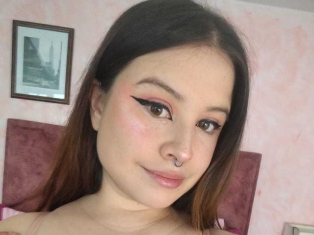 MelaniBecker has brown eyes, black eyeliner, pink eyeshadow, a nose ring, and a slight smile. She has light skin and brown hair, with a pinkish bedroom wall behind.