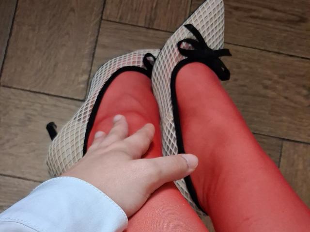 Marriam draagt rode leggings, puntlaarsjes met zwarte strikken en een wit shirt. Ze wordt vastgehouden door een hand op een houten vloer. De borsten zijn niet zichtbaar..