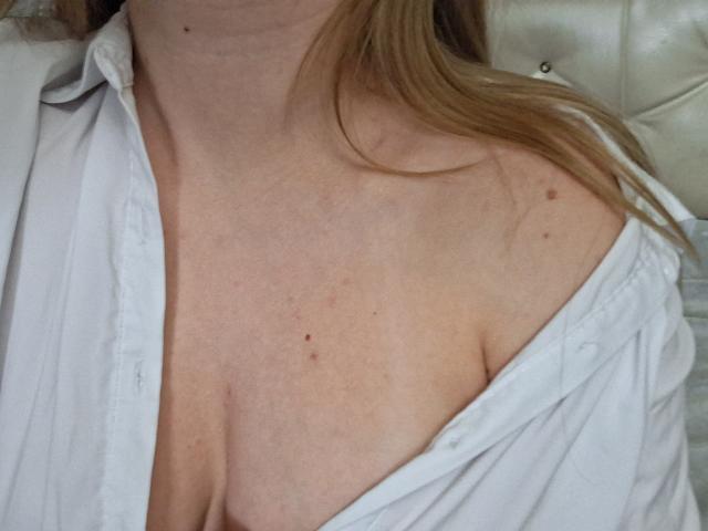 De onbedekte, bleke borst van Marriam, met kleine, donkere moedervlekken en lichte sproeten, draagt een open wit shirt, waardoor haar borsten en een stukje van haar achterwerk zichtbaar zijn. Haar blonde haar is te zien..