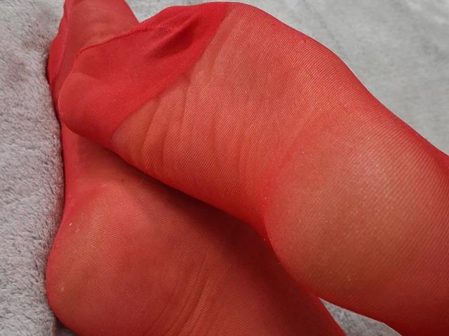 Les orteils rouges et transparents de Marriam sont croisés, révélant le tissu texturé et translucide ainsi que le léger pli de sa peau. Son pied est positionné avec le talon relevé..