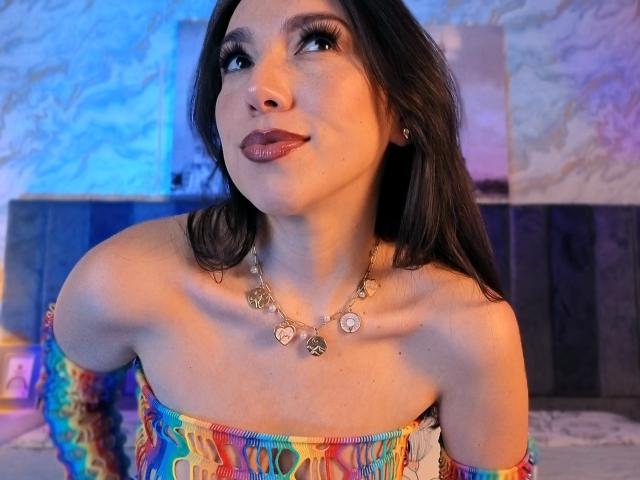 MiaReed with long brown hair, wearing a colorful off-shoulder top and necklace, looking up with a slight smile, showing her tits and a cute face.