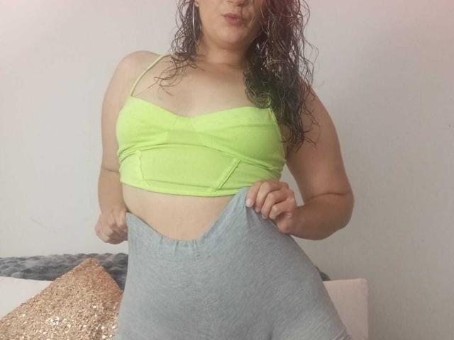 YessiSoto with wet brown hair, green crop top, and gray high-waisted pants, pulling pants up, showing her belly and small tits. She's standing in a room.
