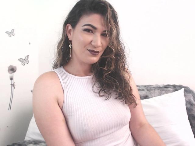 YessiSoto with curly brown hair, white ribbed tank top, dark lipstick, earrings, and a slight smile, showing her tits and ass, sitting on a bed.