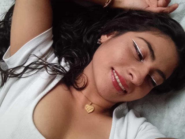 AngelAlba with curly black hair, white tee, and gold necklace, smiling with eyes closed, wearing white eyeliner, lying down, arms above head, visible boobs.