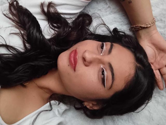 AngelAlba has glossy red lips, dark wavy hair, and white eyeliner, lying on a white surface with a gold bracelet on her wrist, looking seductive.
