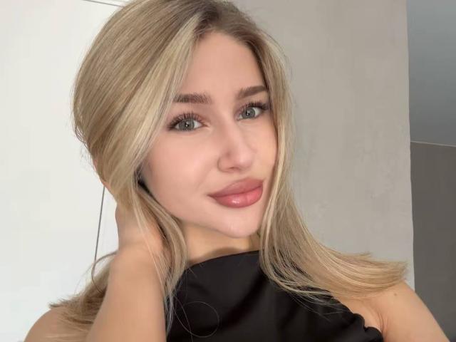 Milagros Edmisten a les cheveux blonds, les yeux verts et porte du rouge à lèvres rose. Elle sourit, une main dans les cheveux, elle porte un haut noir et regarde directement l'appareil photo..