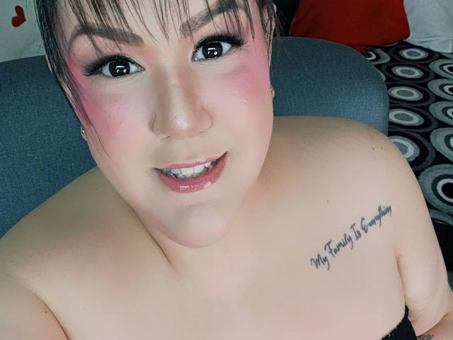 GiiannaMorrys has pink blush on her cheeks, black eyeliner, and a tattoo on her right shoulder saying "My Family is Everything." She's smiling with shiny pink lips.