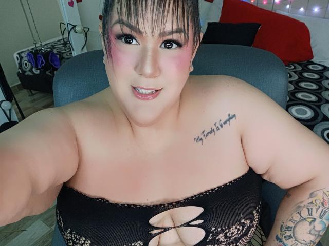 GiiannaMorrys, plus-sized with pink blush on cheeks, black lace top, showing tits, tattoo on right shoulder, arm tattoo, and smiling at camera. Brown hair in ponytail.